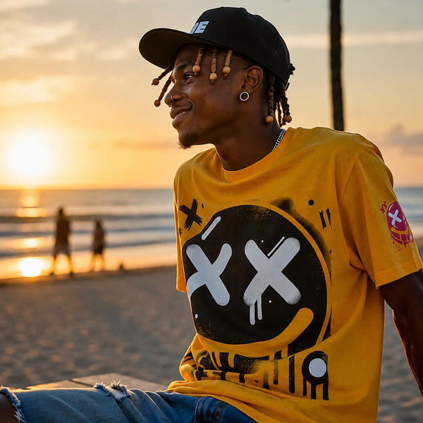 A man wearing a bright yellow CaliPride Clothing graphic t-shirt with  a black cap, and blue jeans, sitting on a beach at sunset.